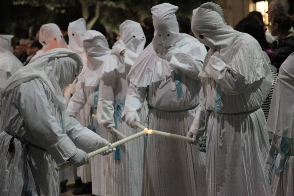 Gruppo serrato di incappucciati in saio bianco con cordone azzurro che si passano la fiamma delle candele