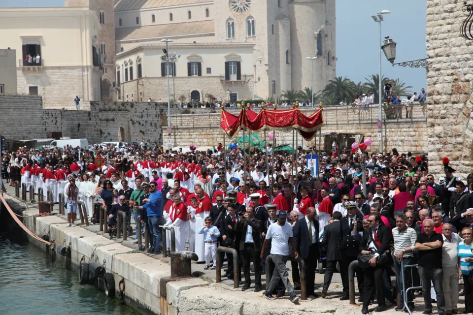 Confraternite in mozzetta rossa, clero e autorità schierati sulla banchina del porto davanti agli edifici in pietra, in attesa del Crocifisso