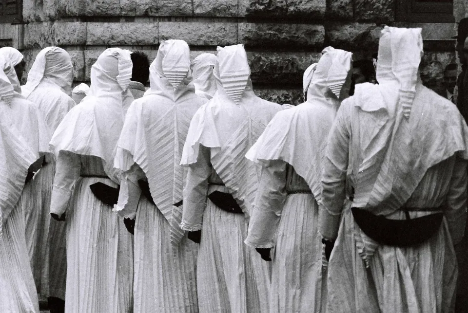 Gruppo di consorelle con veli e cappe bianche e cintura scura, viste di spalle davanti a un muro in conci di pietra
