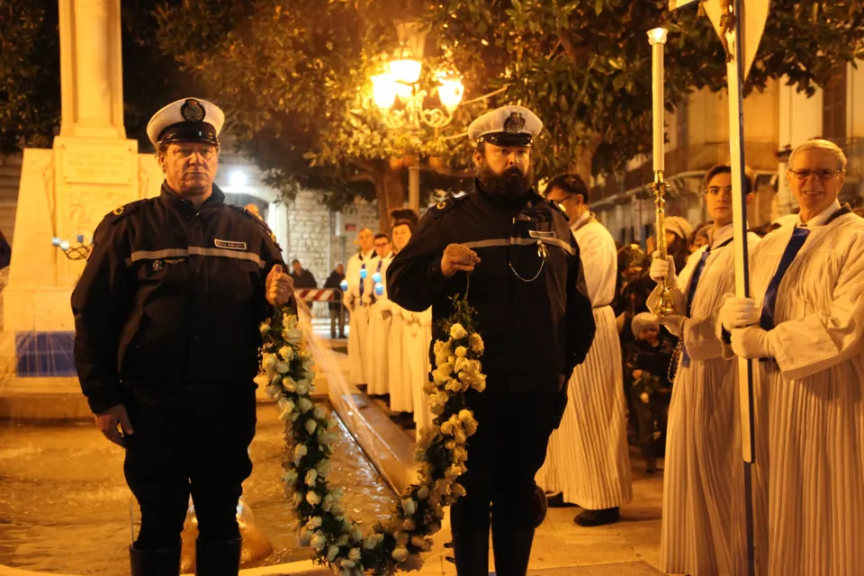 Due agenti della Polizia Municipale reggono la corona di fiori accanto ai chierici con la croce processionale