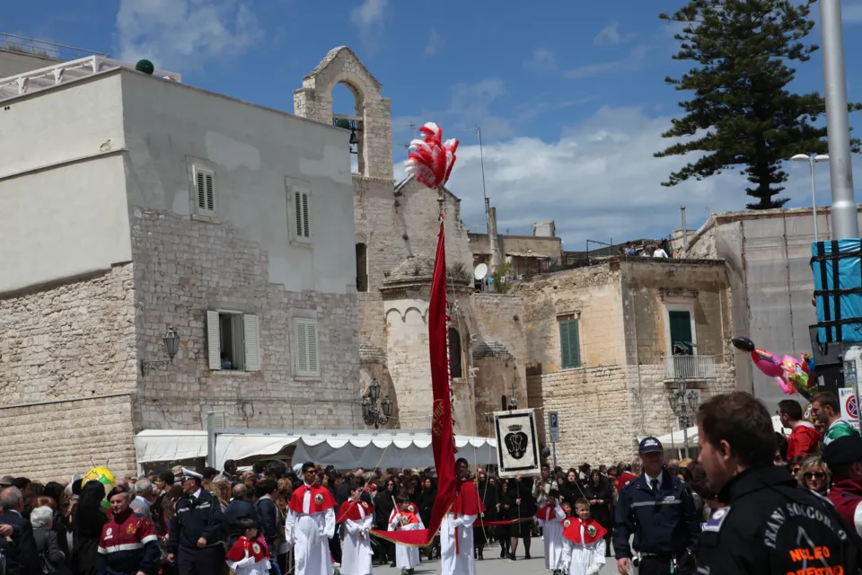 Confratelli con stendardo rosso e fedeli in corteo davanti a edifici in pietra, tra cui una chiesa medievale con campanile a vela