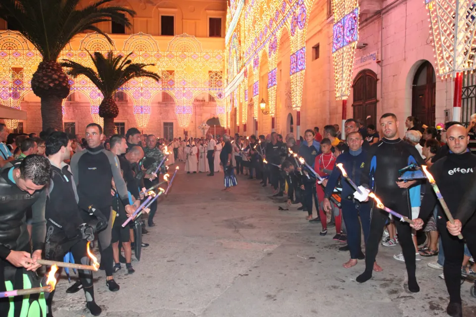 Sommozzatori in muta con fiaccole schierati ai lati della strada sotto le luminarie, con fedeli in attesa del passaggio della processione