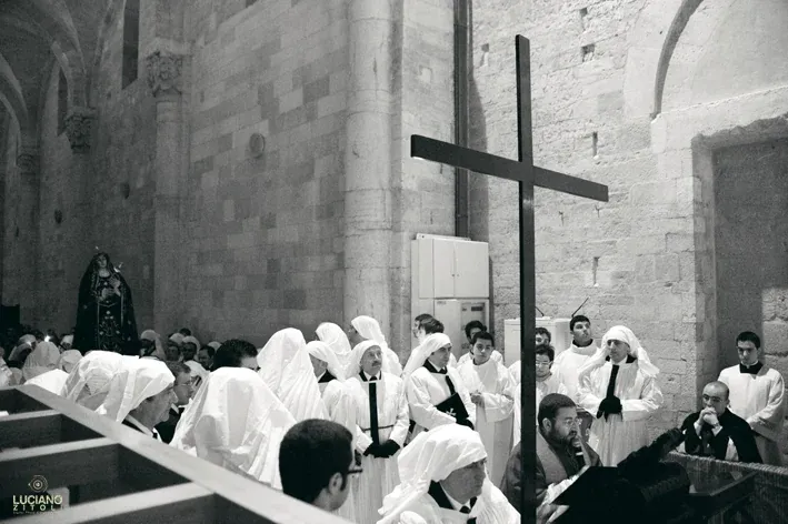 Una grande croce di legno tra confratelli in tunica bianca e fedeli, accanto alla parete in pietra della Cattedrale