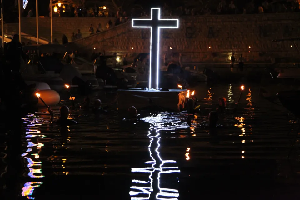 Croce luminosa sostenuta dai sommozzatori nell'acqua del porto, con il riflesso sull'acqua e la banchina sullo sfondo