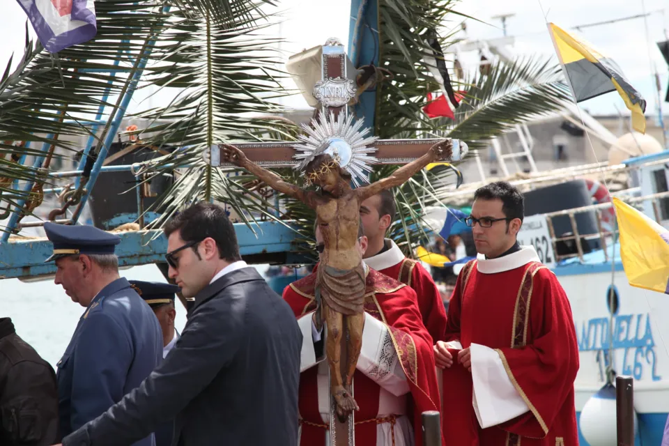 Il Crocifisso di Colonna con raggiera a bordo di un'imbarcazione decorata con palme, tra sacerdoti in rosso e un ufficiale in uniforme