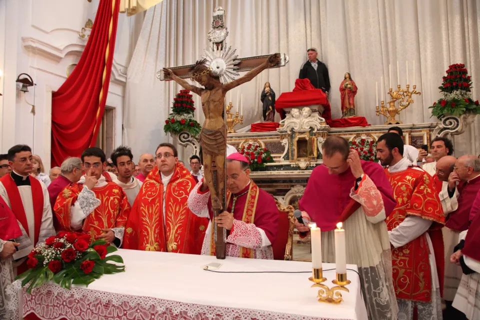Interno di chiesa: il Crocifisso sull'altare mentre un presule e sacerdoti in paramenti rossi celebrano, con fiori rossi e candele