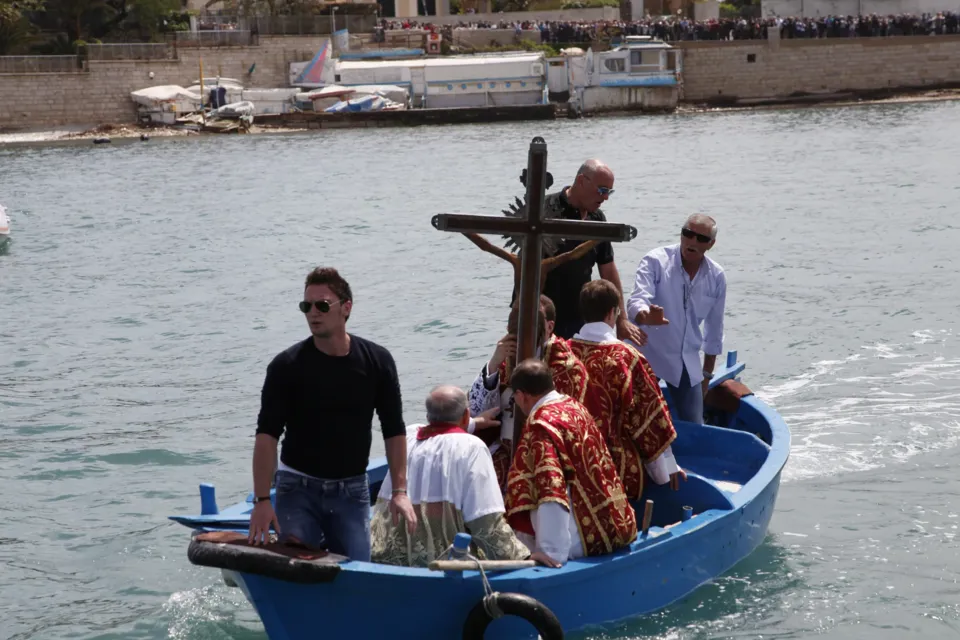 Il Crocifisso di Colonna a bordo di una barca blu in mare, con sacerdoti in paramenti rossi e un uomo in piedi a prua
