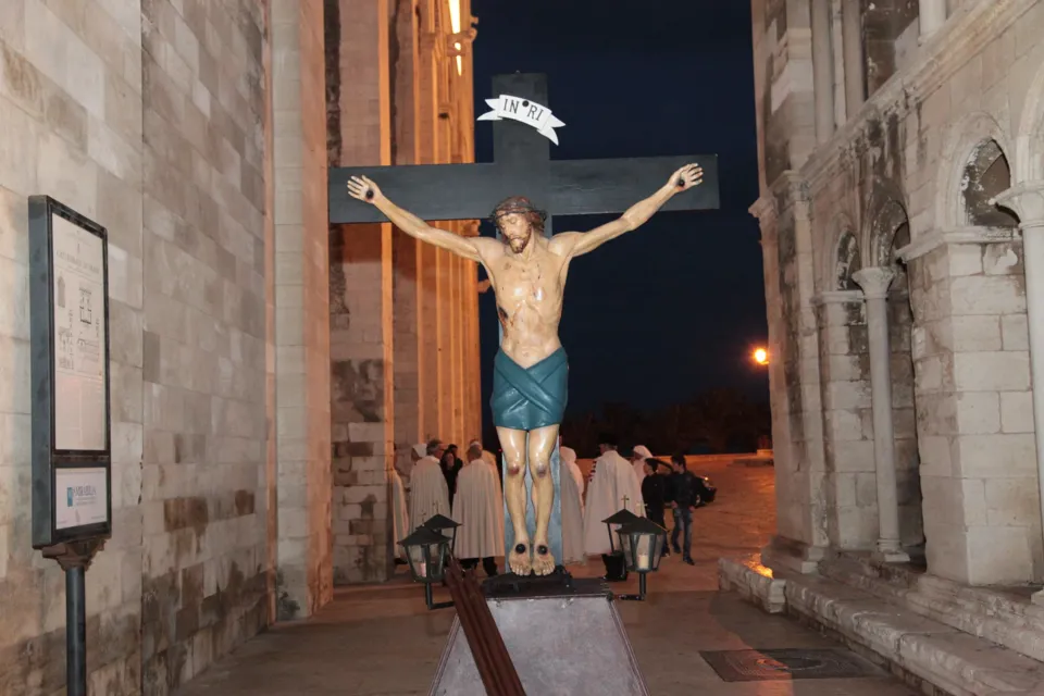 Un grande crocifisso processionale avanza di sera in una stretta via tra edifici in pietra, seguito da confratelli in bianco