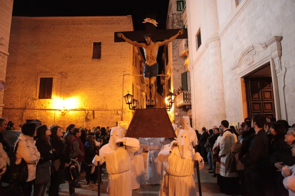 Il grande crocifisso dei Misteri, portato da confratelli incappucciati, attraversa una piazza illuminata gremita di fedeli, accanto alla facciata chiara di una chiesa