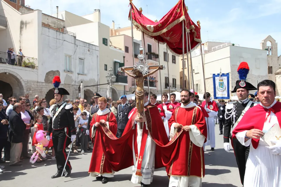 Il Crocifisso sotto il baldacchino rosso portato in processione fra case chiare, con sacerdoti in rosso e carabinieri in alta uniforme