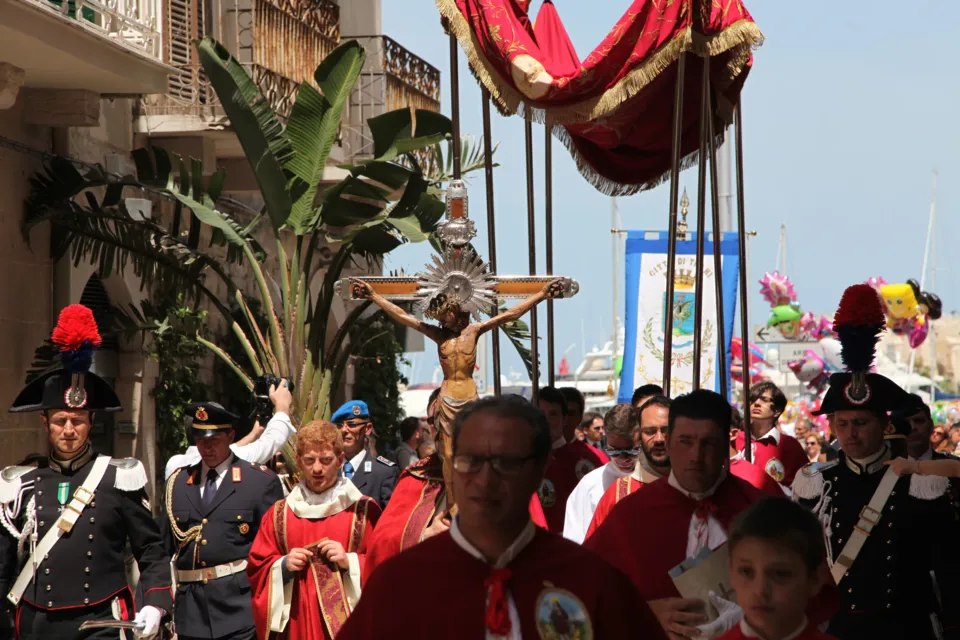 Il Crocifisso sotto il baldacchino rosso in processione tra carabinieri in alta uniforme, confratelli e fedeli, in una via con piante