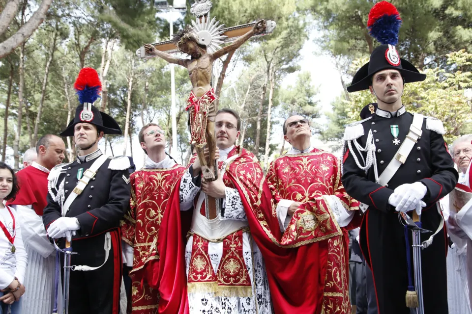 Il Crocifisso portato in processione sotto i pini, tra carabinieri in alta uniforme con pennacchio e sacerdoti in paramenti rossi