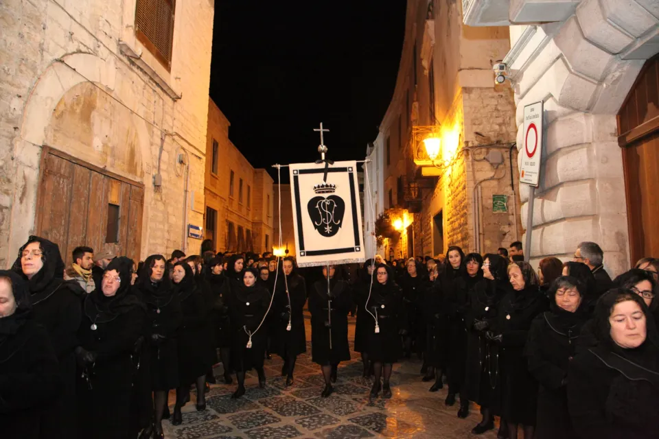 Un lungo gruppo di donne vestite di nero con velo segue uno stendardo nero e bianco in una via del centro storico di notte