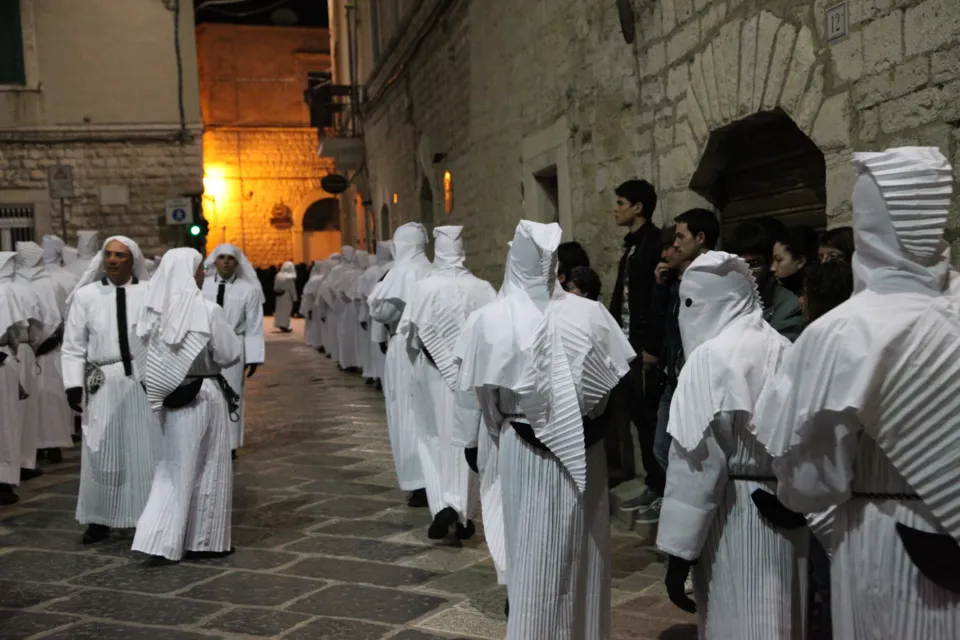 Due file di confratelli incappucciati procedono su una via lastricata del centro storico, tra edifici in pietra e alcuni spettatori
