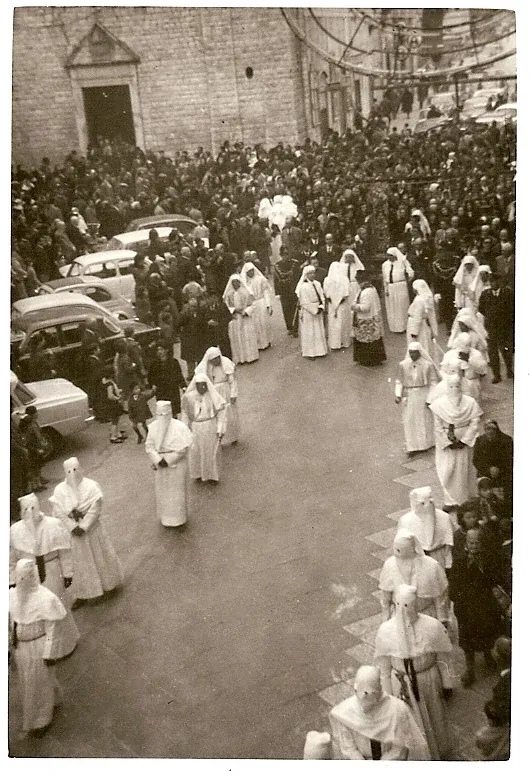 Foto d'epoca di due lunghe file di confratelli in tunica bianca che procedono in una via gremita, con automobili d'epoca a sinistra