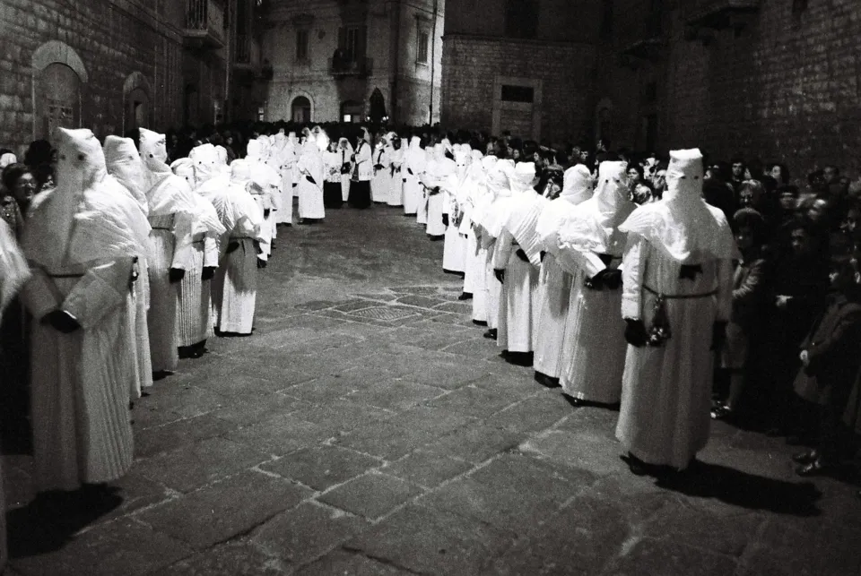 Lunghe file di confratelli con tunica e cappuccio bianchi che si curvano su una strada lastricata, tra edifici in pietra e la folla ai lati