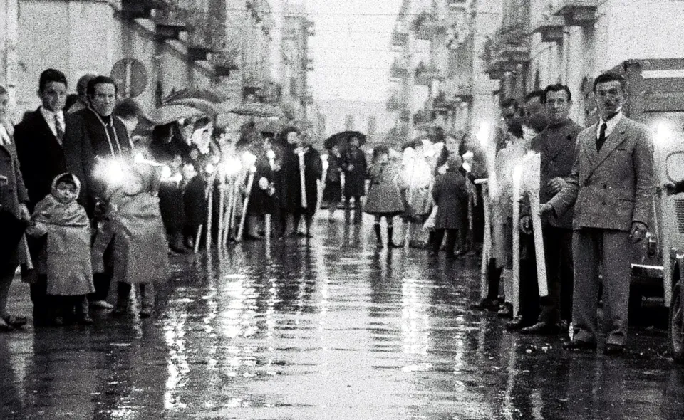 Prospettiva di una via cittadina lucida di pioggia, con due ali di fedeli che reggono ceri accesi in attesa della processione