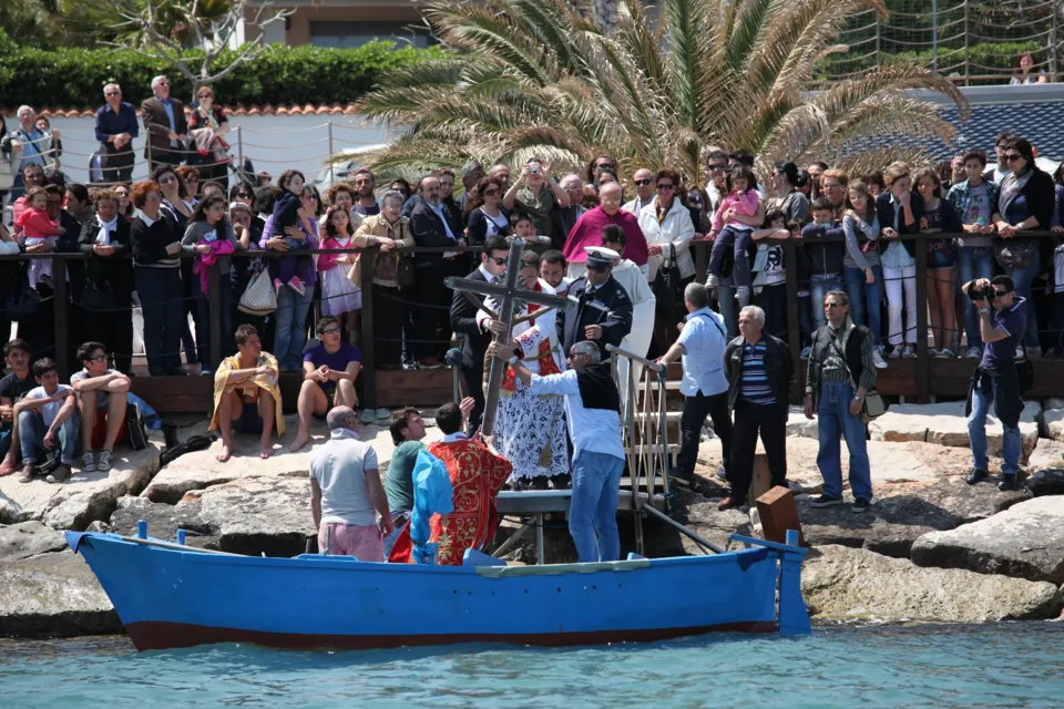 Folla di fedeli sugli scogli e sul molo a Colonna guarda la barca blu su cui viene caricato il Crocifisso