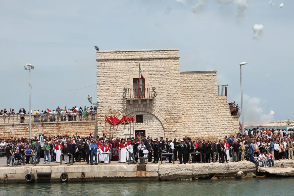 Fortino in pietra decorato con drappi rossi e folla schierata sulla banchina del porto per l'approdo del Crocifisso; fumo di fuochi diurni nel cielo