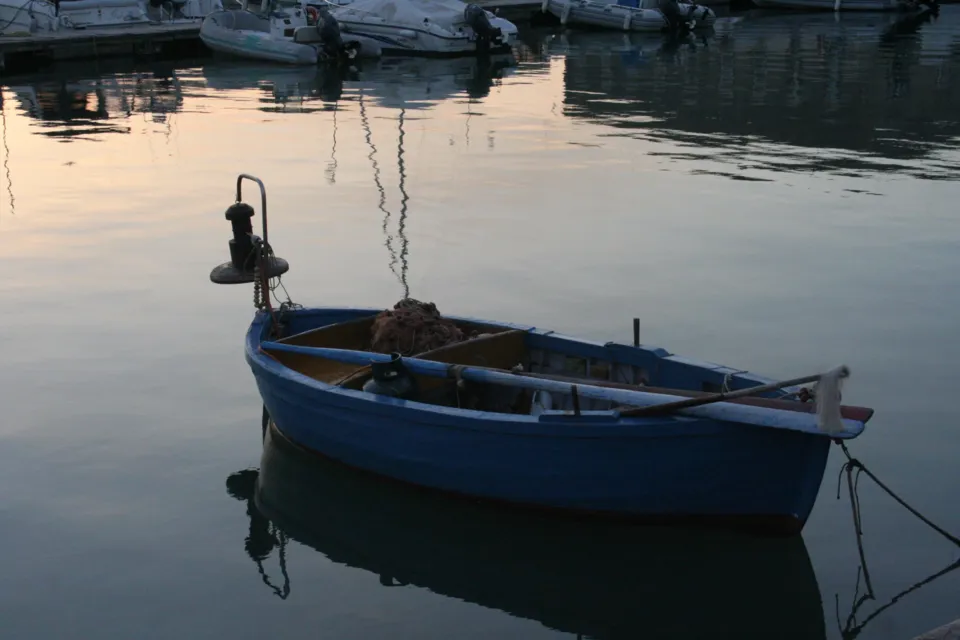Una piccola barca blu con lanterna ormeggiata sull'acqua calma del porto, con riflessi caldi dell'alba