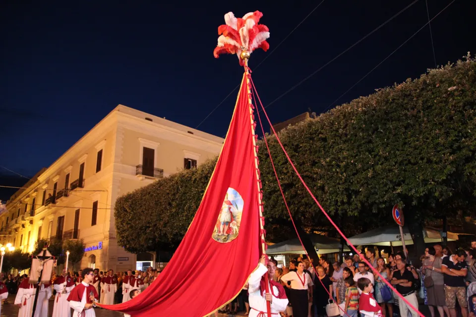 Un grande stendardo rosso con pennacchio, sorretto da funi, avanza di sera lungo una via alberata durante la processione di San Nicola Pellegrino
