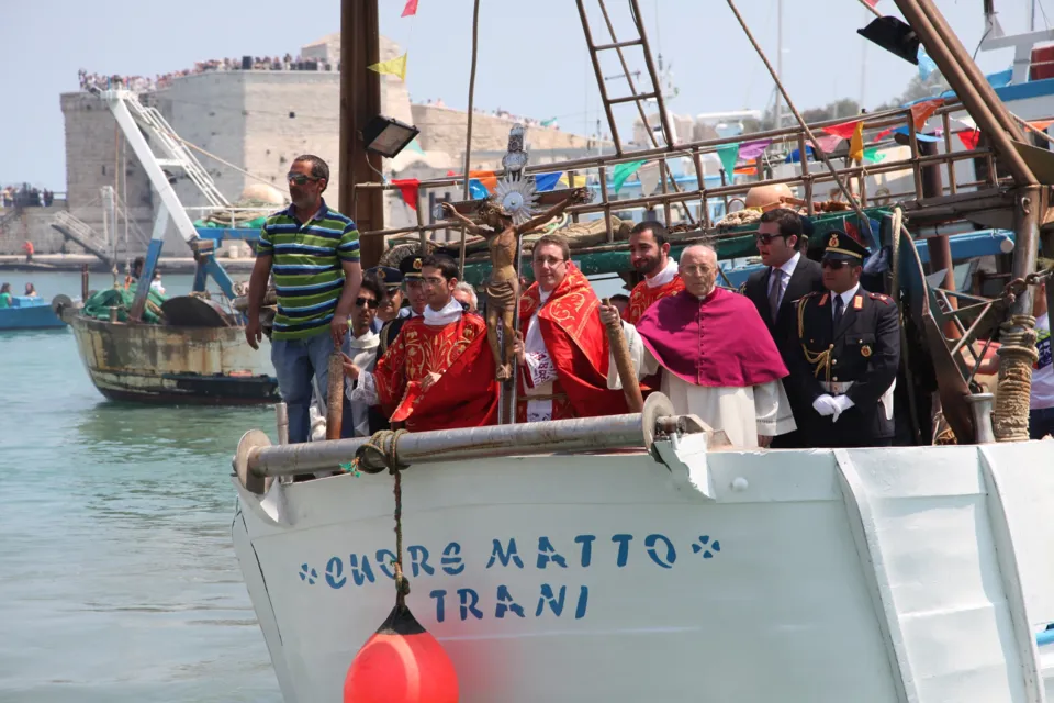 Il Crocifisso a bordo del peschereccio "Cuore Matto" con sacerdoti, un prelato e un agente in uniforme, nel porto con un fortino in pietra sullo sfondo