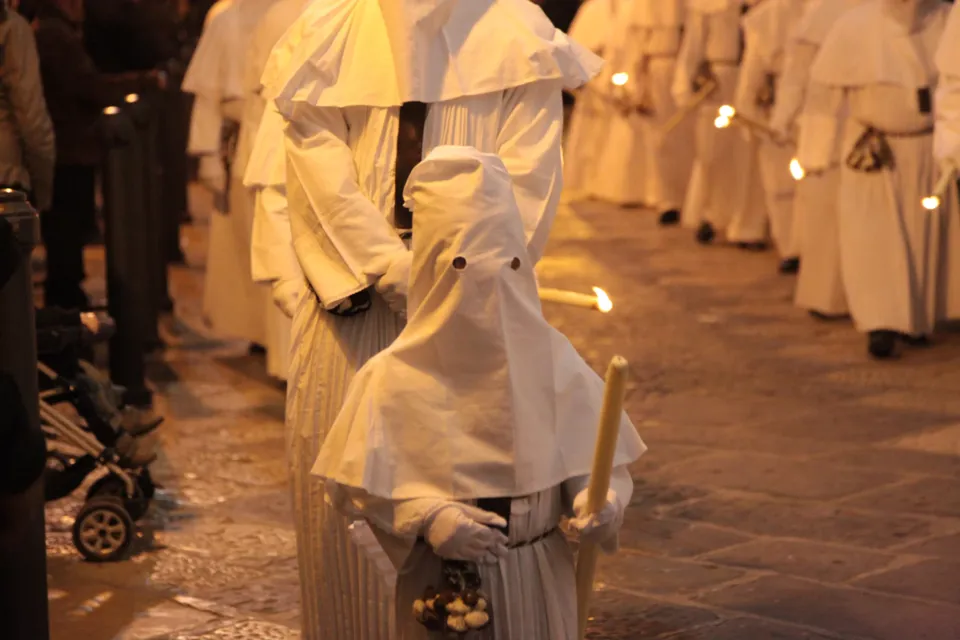 Due file di confratelli in cappuccio bianco con candele accese avanzano su un selciato lucido; in primo piano un incappucciato di spalle