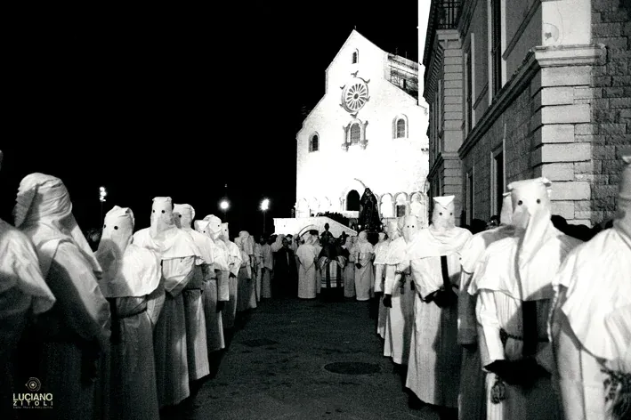 Due file di confratelli incappucciati in bianco e, sullo sfondo, la facciata illuminata della Cattedrale con il rosone, di notte