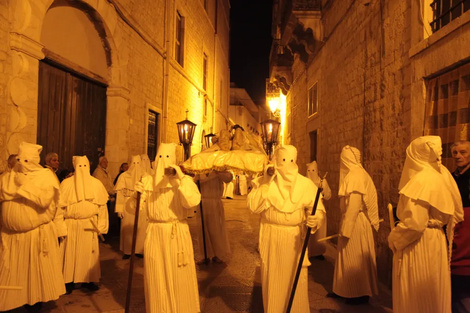 Confratelli con cappuccio bianco a punta e bastone avanzano in un vicolo del centro storico illuminato da luce calda