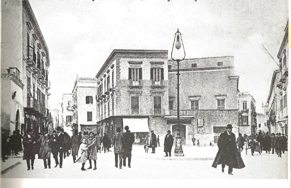 Fotografia d'epoca di un incrocio cittadino con palazzi, passanti e un alto lampione al centro