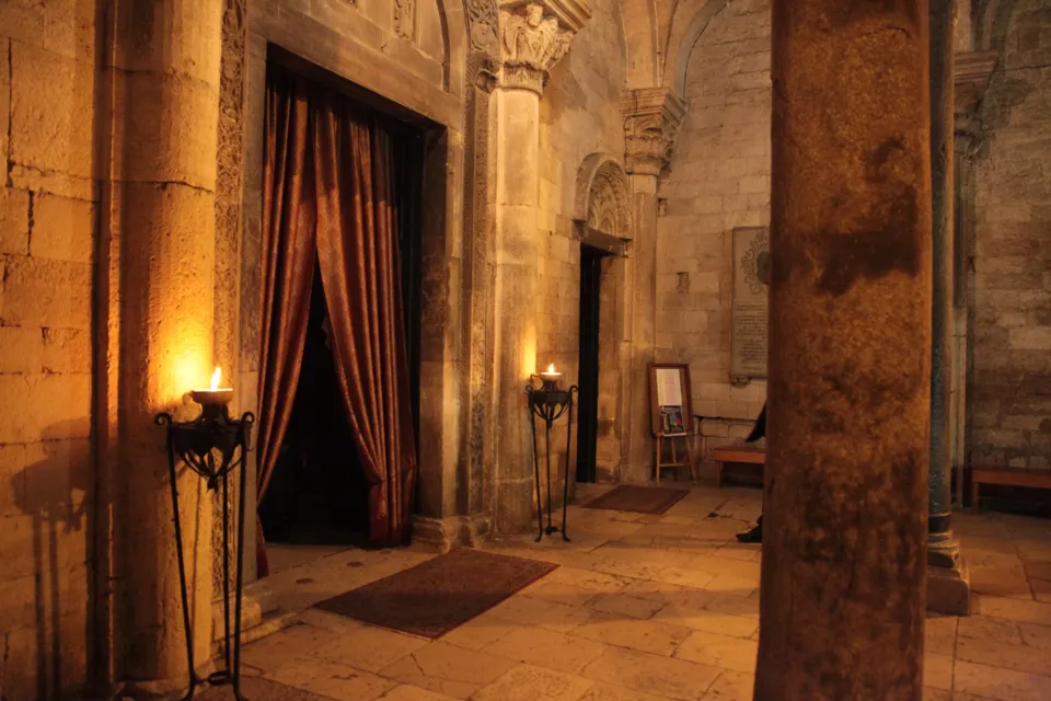 Interno di una chiesa romanica in penombra con colonne, capitelli scolpiti, una porta chiusa da tende rosse e bracieri accesi