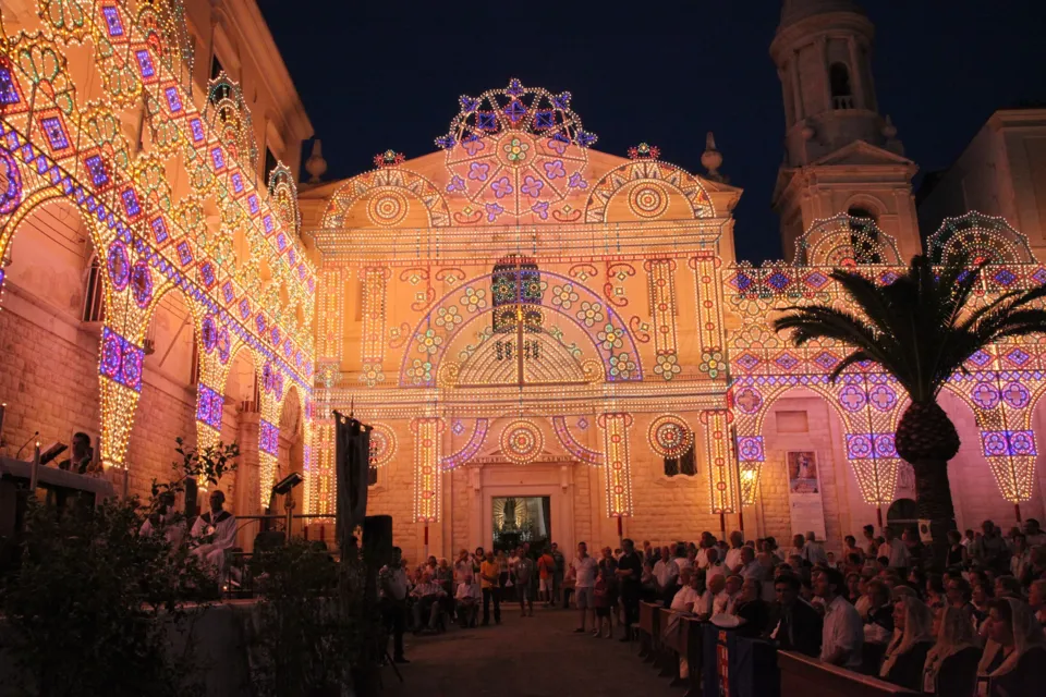 Grande facciata di luminarie colorate allestita davanti alla chiesa per la festa della Madonna del Carmine, con folla nella piazza