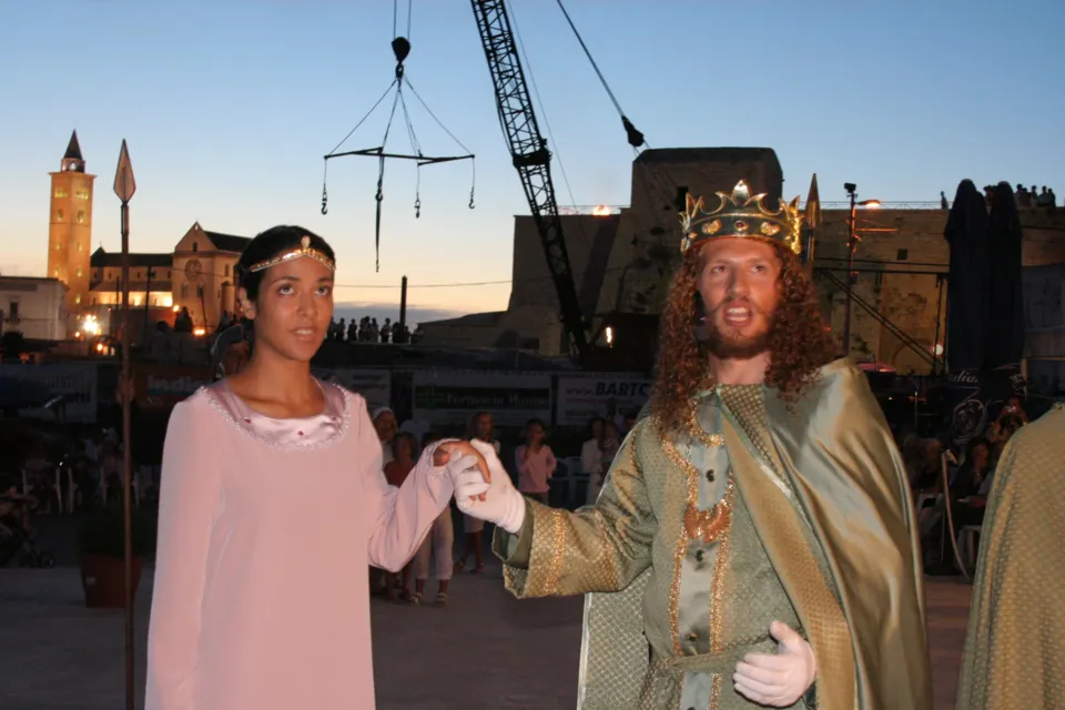 La coppia reale in costume, mano nella mano, sul porto al crepuscolo; sullo sfondo il campanile della Cattedrale e il Castello illuminati e una gru