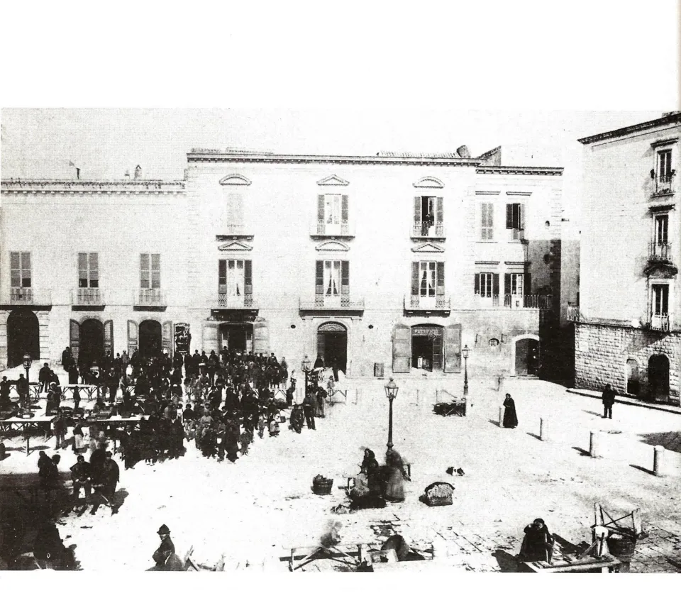 Fotografia d'epoca di una piazza con venditori e banchi del mercato davanti a palazzi ottocenteschi, con un lampione al centro