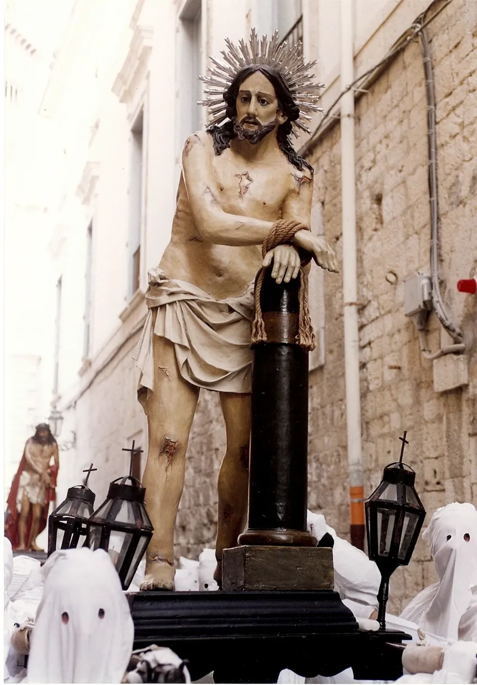 Statua del Cristo legato alla colonna, con aureola raggiata, portata su base con lanterne tra confratelli incappucciati