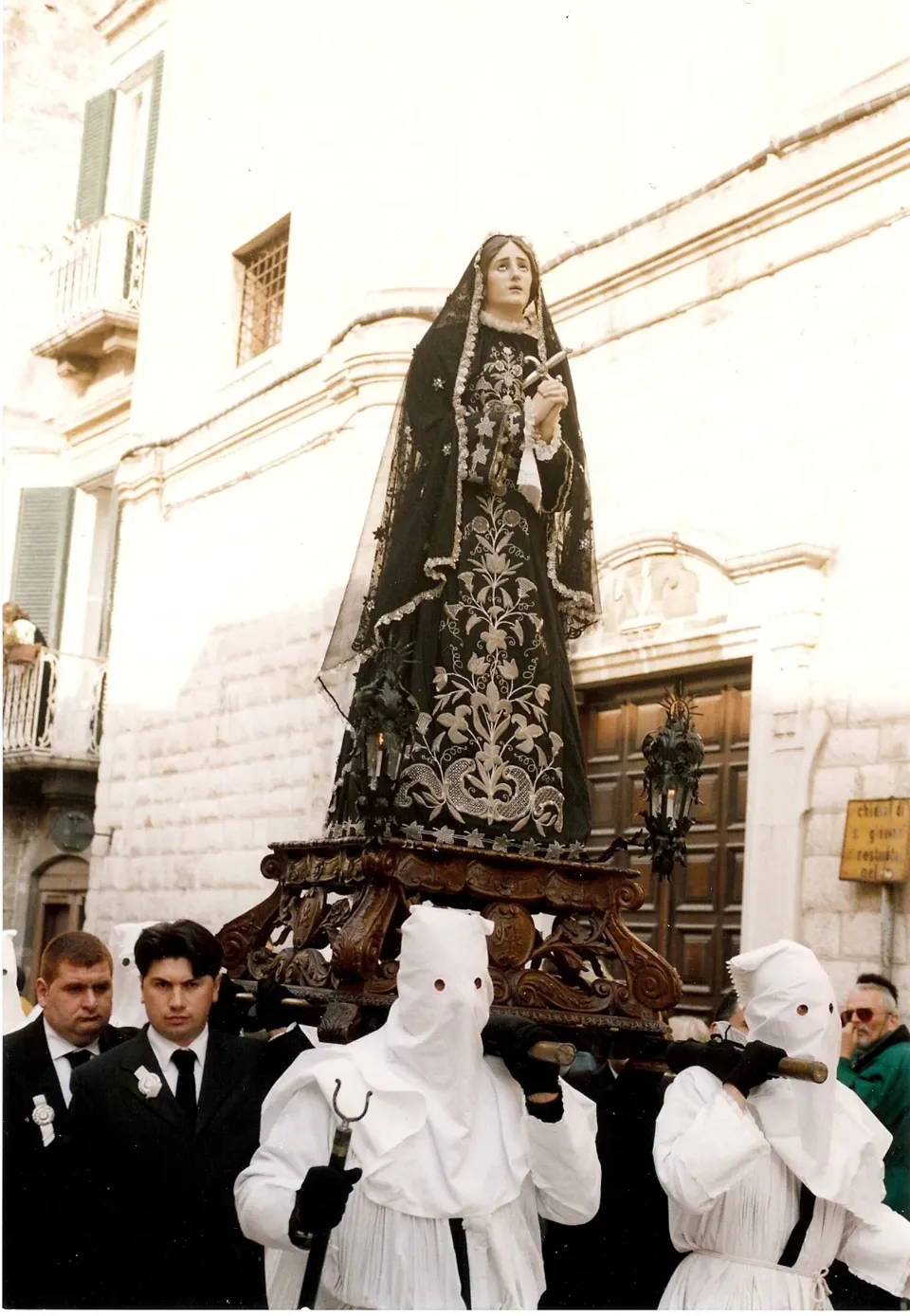 Statua dell'Addolorata con manto nero ricamato portata in processione da uomini in abito scuro e da confratelli incappucciati in bianco