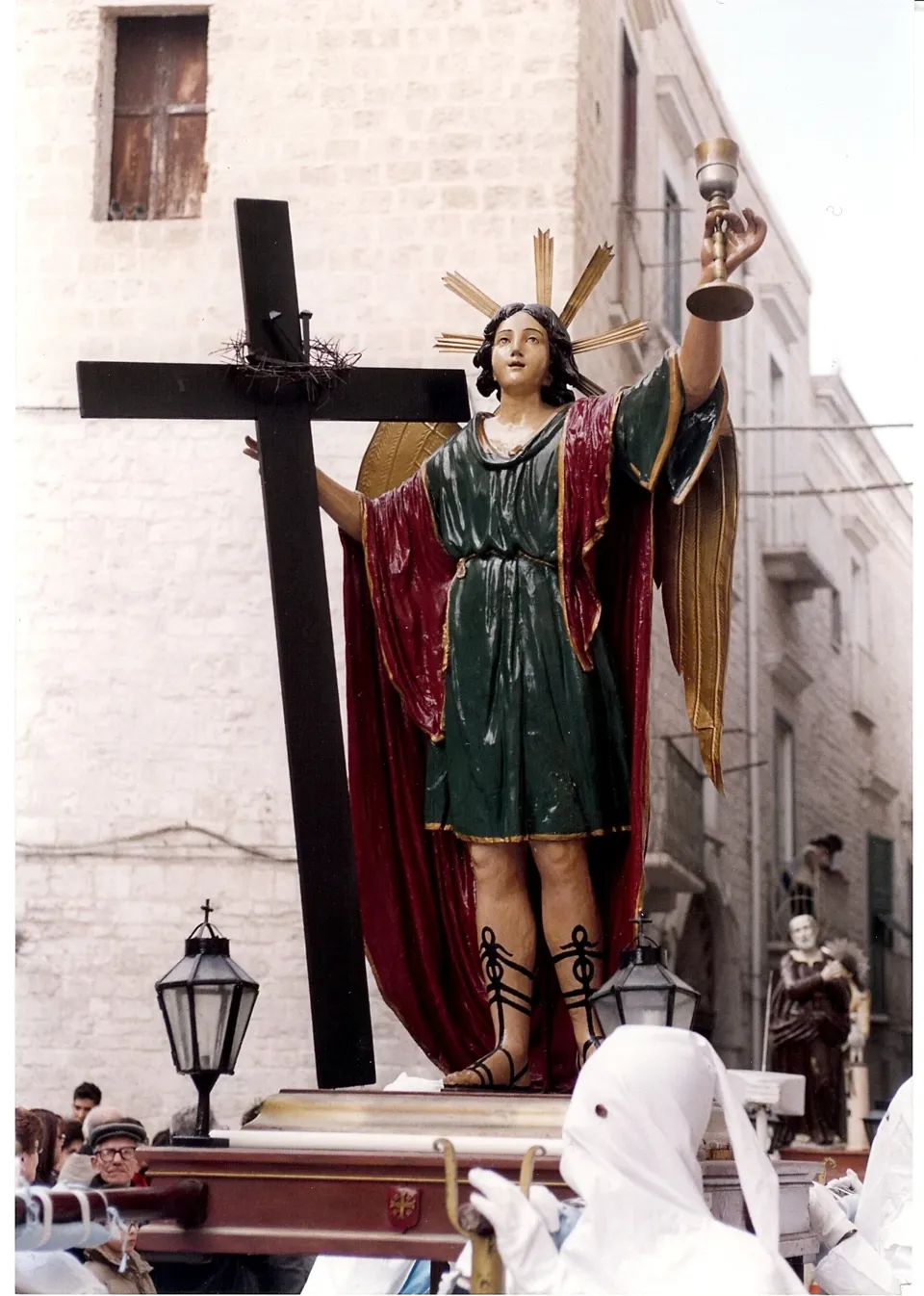 Statua di un angelo con ali dorate che regge un calice, accanto a una grande croce scura, portata in processione