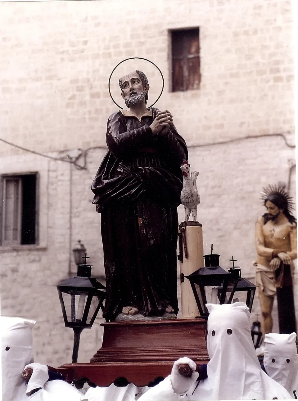 Statua di San Pietro con aureola e mani giunte al petto, portata su base con lanterne da confratelli incappucciati in bianco, tra le case in pietra chiara