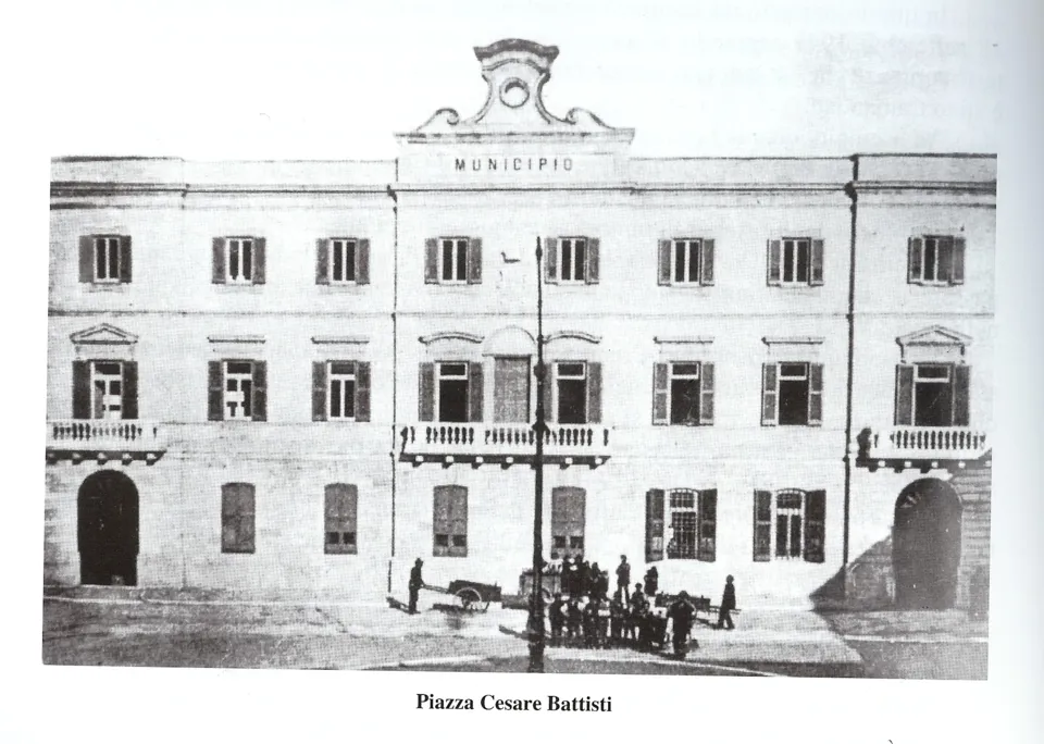 Foto d'epoca del palazzo del Municipio con alcune persone davanti, didascalia 'Piazza Cesare Battisti'