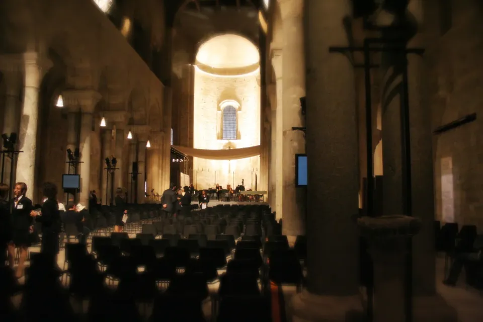 Interno della Cattedrale in penombra con file di sedie nella navata, colonne e abside illuminata