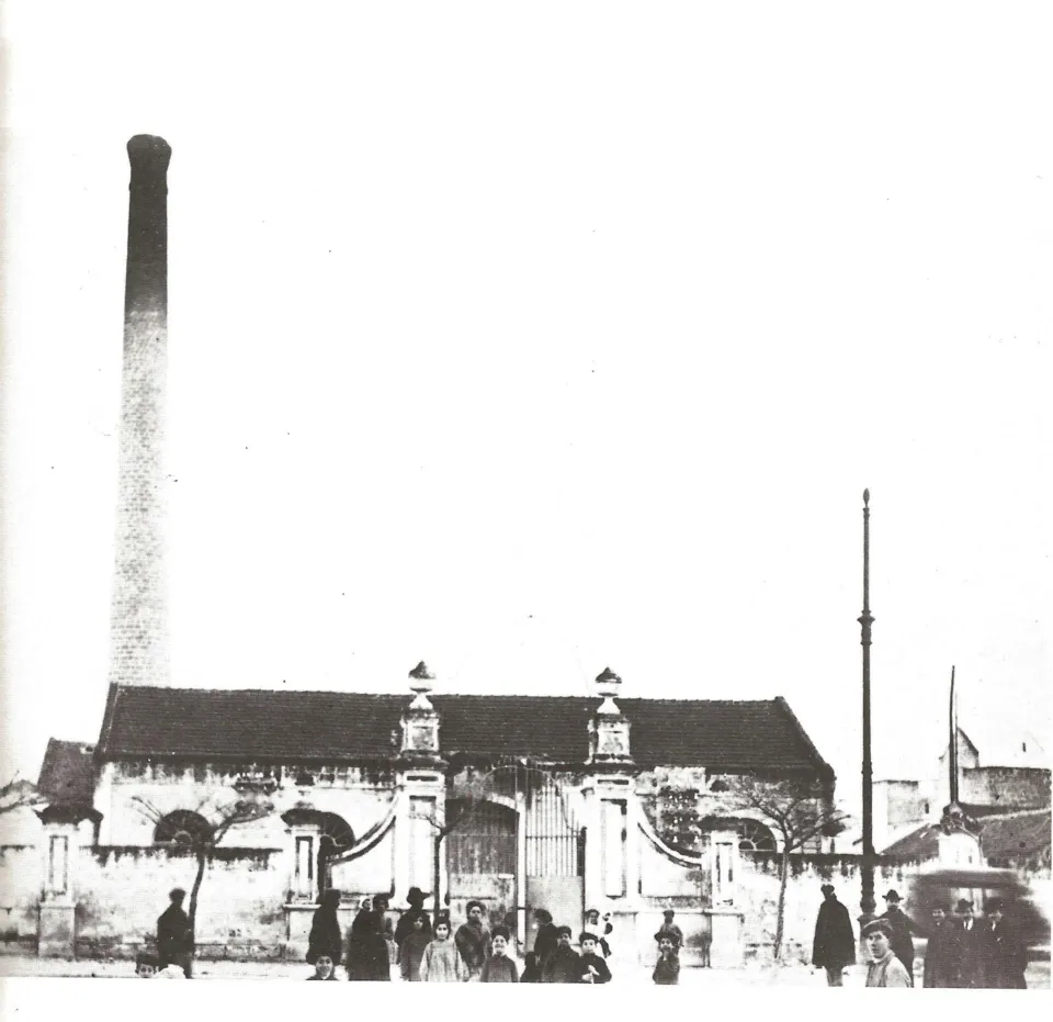 Fotografia d'epoca di un edificio industriale con alta ciminiera in mattoni e cancello ornato, con alcune persone davanti