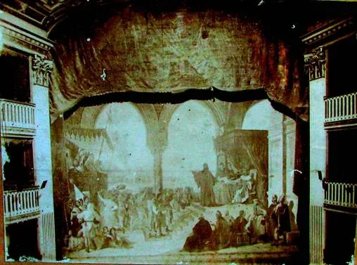 Foto d'epoca del boccascena del Teatro comunale di Trani con i palchi laterali e il sipario dipinto calato, raffigurante una scena medievale sotto una loggia gotica