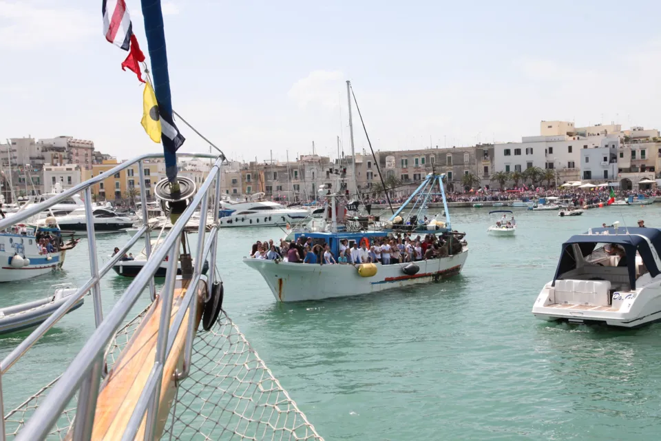 Dalla prua di un veliero: un peschereccio carico di fedeli e motoscafi nel porto, con le case del lungomare sullo sfondo
