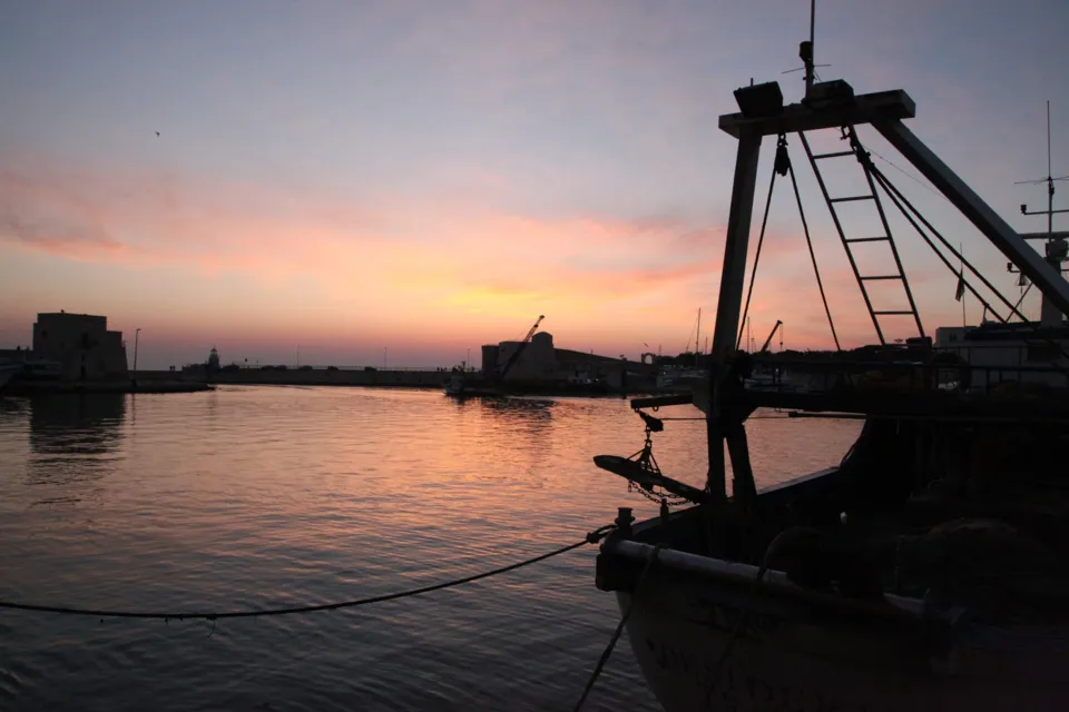 Sagoma di un peschereccio ormeggiato in controluce sul bacino del porto, con il cielo rosa dell'alba e il profilo del fortino sullo sfondo