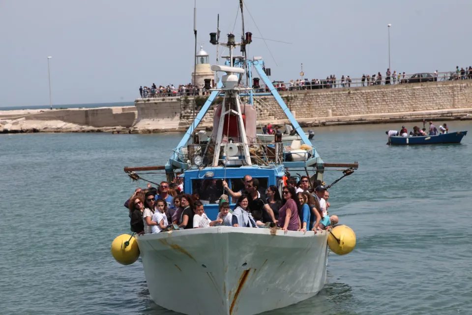 Peschereccio con fedeli a bordo che entra nel porto, con il molo affollato e una barca a remi sullo sfondo