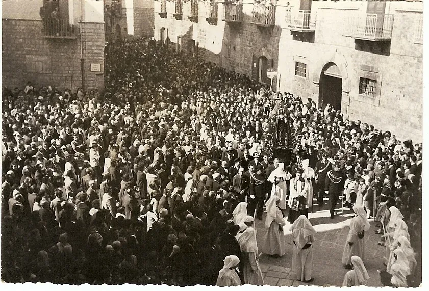 Foto d'epoca di una piazza gremita di fedeli, con confratelli in bianco e una statua al centro, tra palazzi con balconi