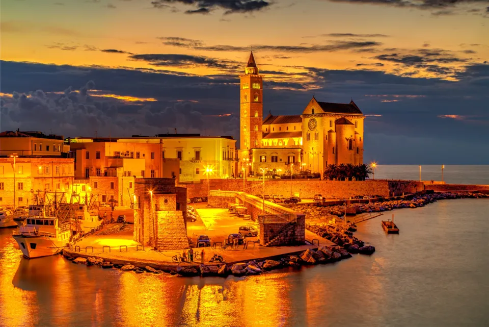 Il porto di Trani al crepuscolo con barche ormeggiate, il lungomare illuminato e, sullo sfondo, la Cattedrale e il campanile illuminati sotto un cielo con nuvole arancioni