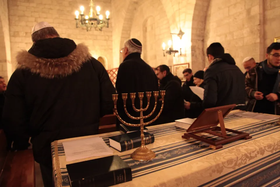 Fedeli con kippah in preghiera nella sinagoga; in primo piano una menorah dorata e libri sul tavolo, in alto un lampadario
