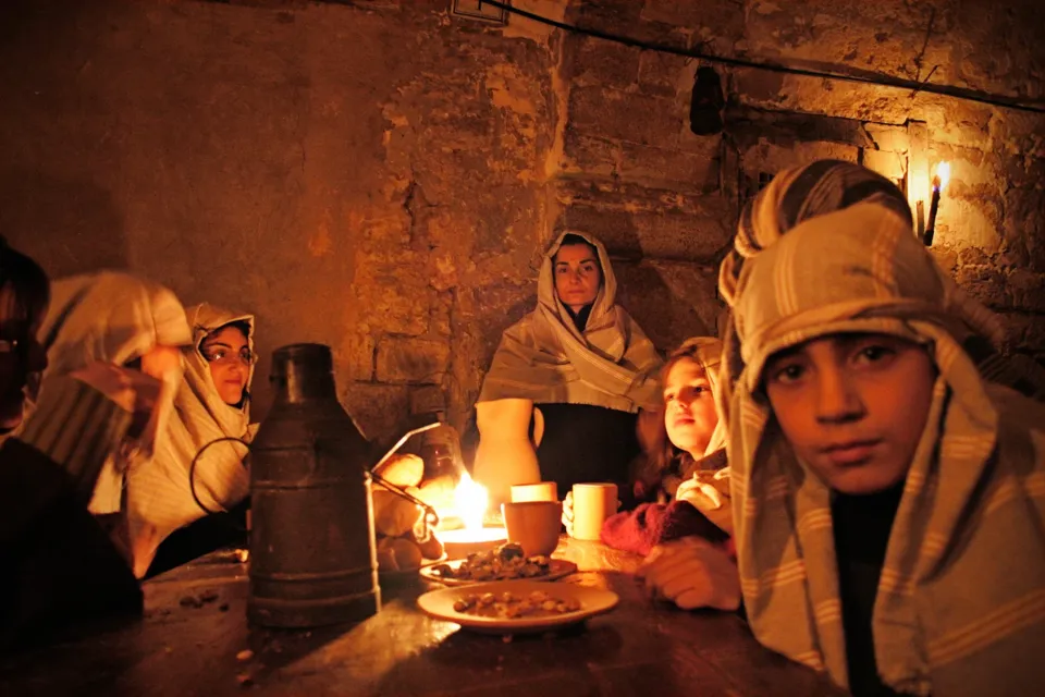 Figuranti in costume, tra cui bambini, attorno a un tavolo illuminato da una candela in un ambiente in pietra