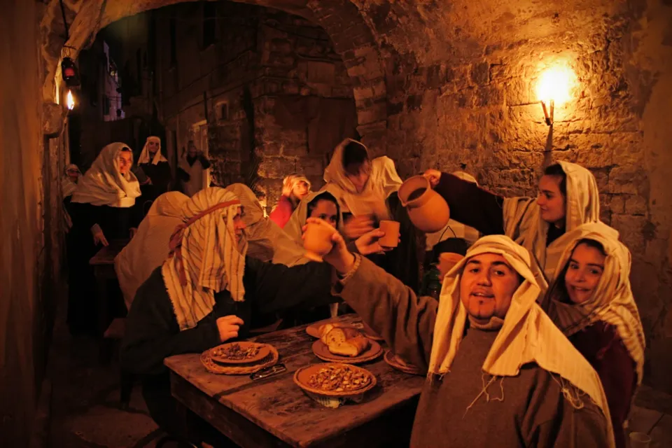 Figuranti in costume con veli e mantelli attorno a un tavolo con cibi, in un ambiente in pietra illuminato da una lanterna